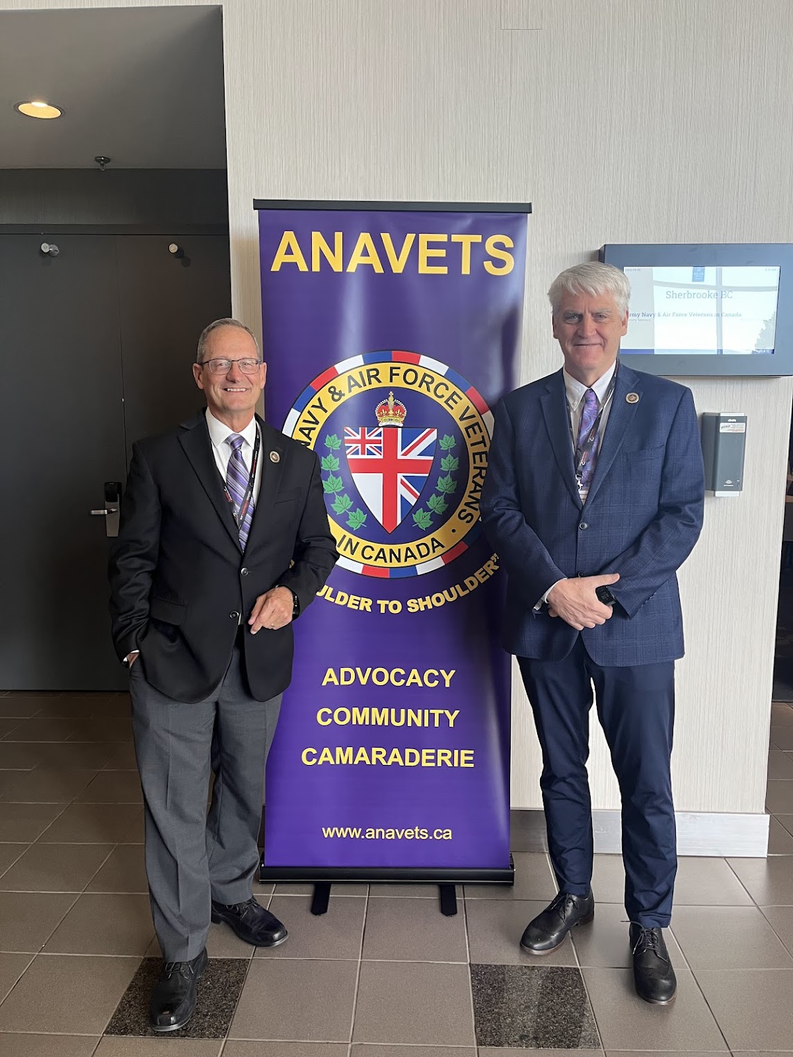 Veterans Review and Appeal Board’s Chairperson Christopher J. McNeil and Deputy Chairperson Jacques Bouchard standing beside a purple banner that reads “ANAVETS – Army, Navy & Airforce Veterans in Canada. Shoulder to shoulder. Advocacy, Community, Camaraderie”.