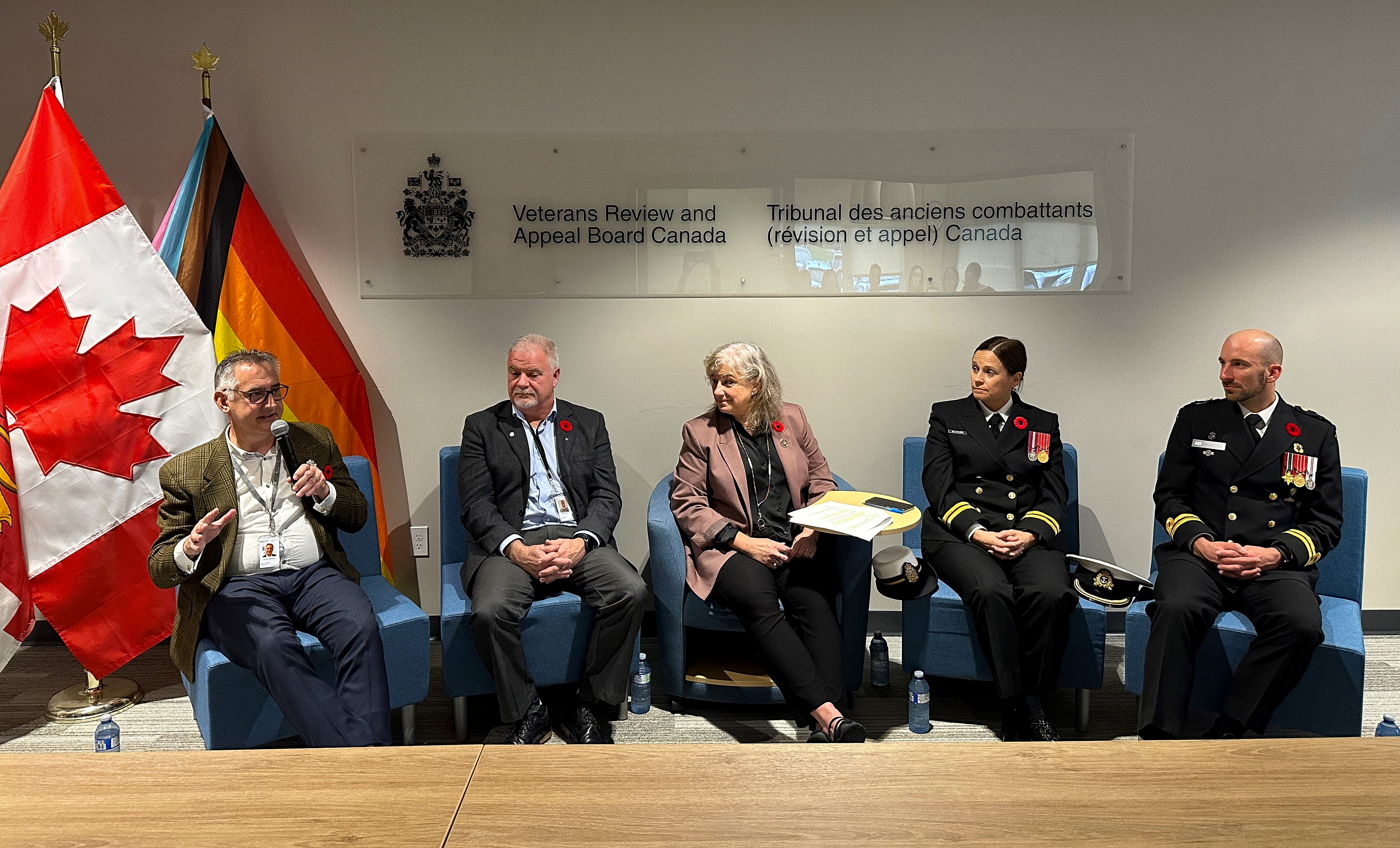 Five individuals sitting on blue chairs in front of the Veterans Review and Appeal Board’s logo and next to a Canadian and a pride flag
